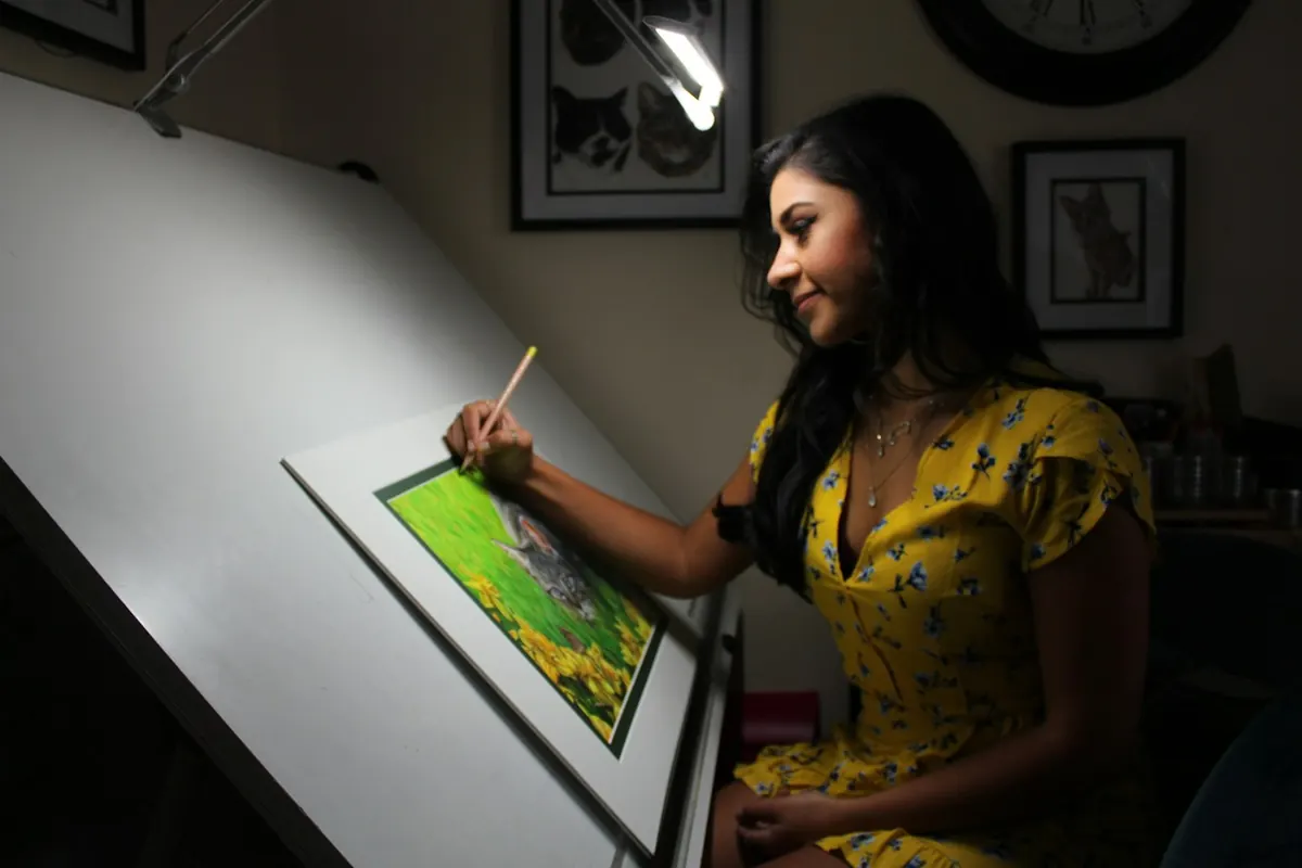 Pessoa a desenhar a lápis sobre uma mesa de desenho inclinada, iluminada por um candeeiro articulado